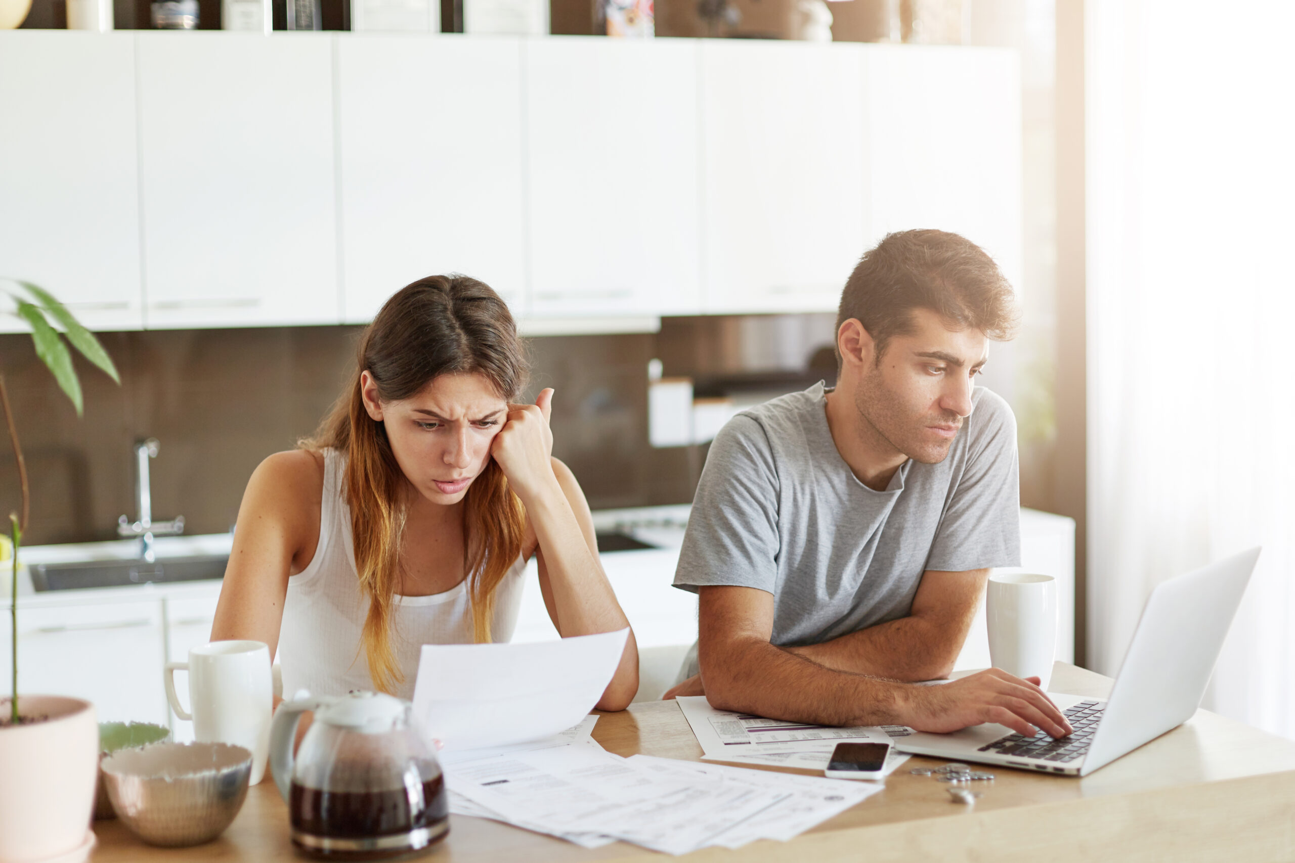 Blog young caucasian couple having concentrated look, doing paper work and working with laptop computer while sitting in kitchen. young family couple managing finances, checking their family budget